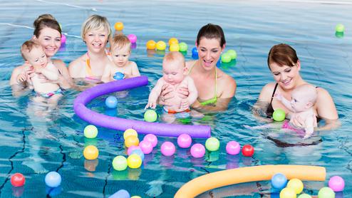 Vier Frauen stehen im Wasser und halten Babys, umgeben von bunten Bällen und Schaumstoffringen in einem Schwimmbad.
