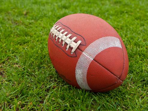 Nahaufnahme eines American-Football auf grünem Gras, im Hintergrund Spieler in gelben Trikots stehend.