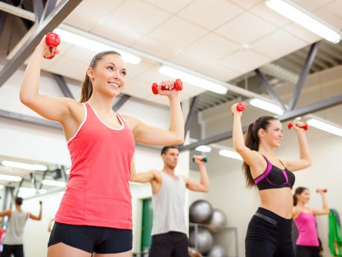 Vier Personen heben Gewichte in einem Fitnessstudio. Zwei Frauen tragen Sporttops, ein Mann hat ein T-Shirt an.
