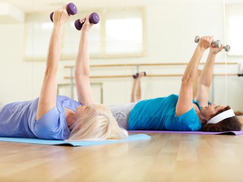 Zwei Frauen liegen auf Gymnastikmatten und heben Gewichte in einer Fitnessumgebung.