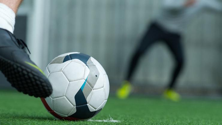 Nahaufnahme eines Fußballes, der auf grünem Kunstrasen vor einem verschwommenen Spieler liegt.