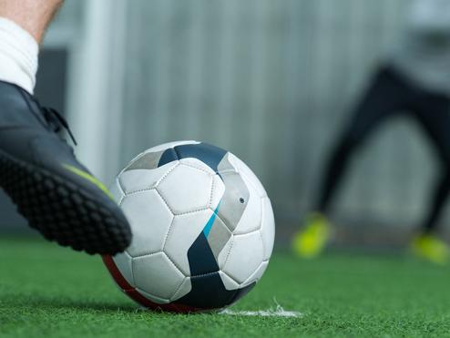 Nahaufnahme eines Fußballes, der auf grünem Kunstrasen vor einem verschwommenen Spieler liegt.