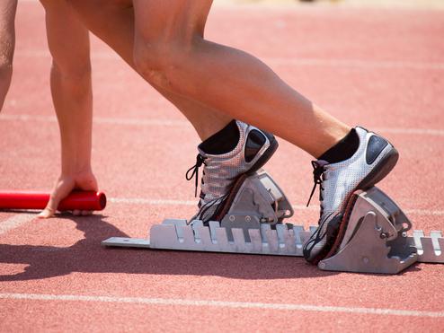 Startposition eines Sprintläufers mit Fuß auf einem Startblock auf einer roten Laufbahn, Hand hält einen Staffelstab.