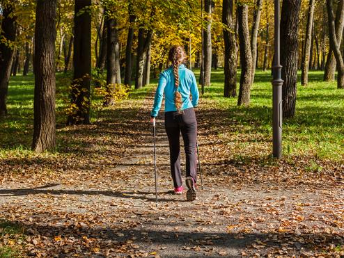 Frau mit langem Zopf geht mit Wanderstöcken einen herbstlichen Weg zwischen Bäumen, umgeben von buntem Laub.